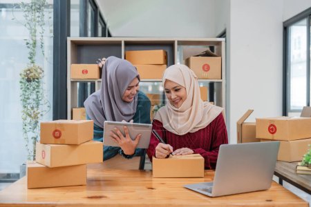 Foto de Muslim dos mujeres freelancer sme negocio de compras en línea de trabajo en el ordenador portátil y lista de verificación caja de pedidos en el hogar Negocios concepto de envío y entrega en línea. - Imagen libre de derechos