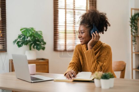 Photo for Smiling young african american woman in wireless headphones sitting at desk table working on laptop and writing letter in paper notebook, taking notes watching weninar. - Royalty Free Image