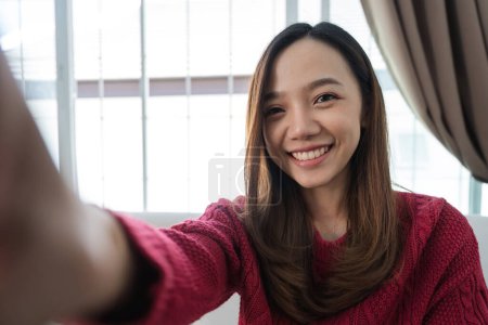 Téléchargez les photos : Jolie jeune femme asiatique avec un grand sourire assis au salon. Elle s'amuse à prendre un selfie joyeux. - en image libre de droit