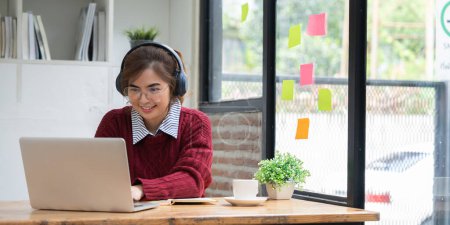 Foto de Asiática estudiante universitaria en casa vistiendo camisa roja usando portátil asistir a clase universitaria en línea escuchando con auriculares. - Imagen libre de derechos