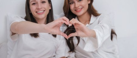 Photo for Lesbian couple together concept. Portrait of a smiling young lesbian couple other while sitting together and hand made heart symbol in the morning at bedroom. - Royalty Free Image