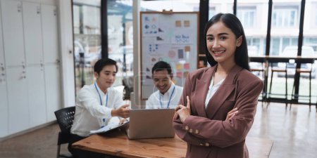 Photo for Portrait of young attractive Asian woman with successful conference room business people in the background while at her office desk smiling - Royalty Free Image
