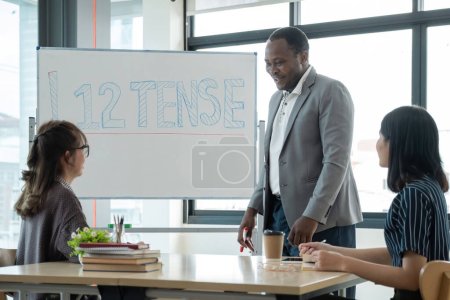 Photo for Positive friendly African American English tutor giving language courses to group of interested adults of different ages and nationalities sitting around table in classroom, Education concept. - Royalty Free Image
