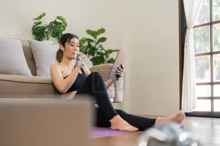 Téléchargez les photos : Femme asiatique jouant tablette à la maison pendant la pause de l'exercice et boire de l'eau douce. - en image libre de droit