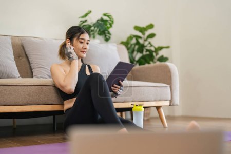 Photo for Lifestyle asian young fitness woman holding tablet relaxing after workout at home. Smiling female using cell phone checking newsfeed on social media while exercise. - Royalty Free Image