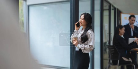 Téléchargez les photos : Femme asiatique parlent au téléphone avec les gens d'affaires de la salle de réunion en arrière-plan tandis qu'à son bureau. - en image libre de droit
