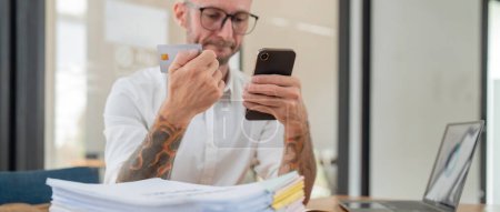 Photo for Stressed caucasian male business holding credit card making online payment using smartphone at office. - Royalty Free Image