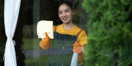 Foto de Concepto de trabajo doméstico y limpieza. mujer feliz en guantes ventana de limpieza con trapo y spray limpiador en casa. - Imagen libre de derechos