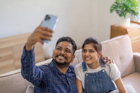 Foto de Feliz lindo joven indio pareja tomando selfie en casa sentado en el sofá. - Imagen libre de derechos
