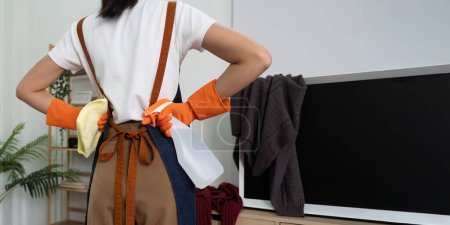 Photo for Tired household clean up, housekeeper asian young woman stand and look many clothes on television, cleaning in living room at home. Mess maid or housewife organize dirty and untidy. - Royalty Free Image