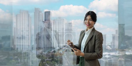 Téléchargez les photos : Jeune asiatique femme exécutif sourire ou professionnel femme d'affaires en costume tenant tablette et debout à côté de la fenêtre avec confiance après le succès. - en image libre de droit
