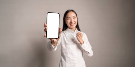 Foto de Mujer asiática alegre, mostrando su teléfono inteligente con pantalla blanca vacía maqueta, se regocija en el éxito, mira a la cámara, hacer el gesto SI. - Imagen libre de derechos