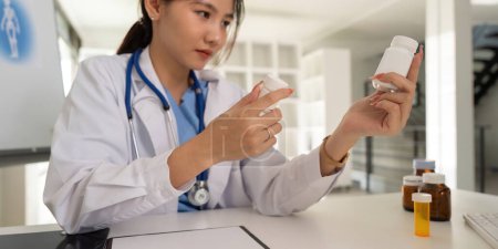 Photo for Female doctor writing in a notebook and holding a medicine bottle works at a computer while the table while sitting at a work desk. - Royalty Free Image