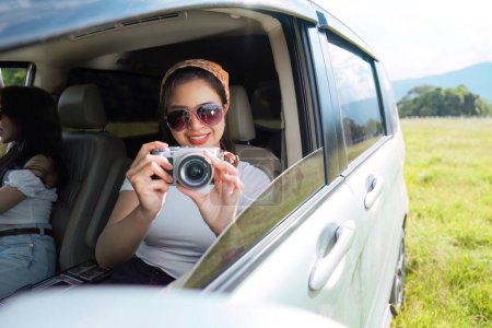 Téléchargez les photos : Belles amies souriantes partent en voyage tout en sortant la tête hors de la voiture en mouvement pour voir la vue et prendre des photos. - en image libre de droit