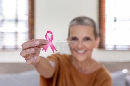 Portrait of confident smiling elderly senior woman with pink ribbon at her home. Health care, support, prevention. Breast cancer awareness month concept.
