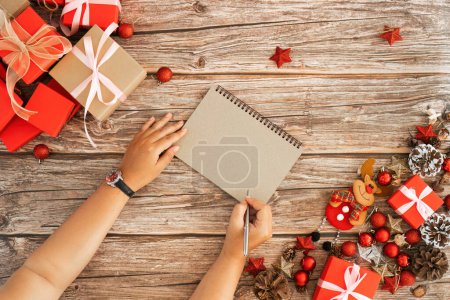 Photo for Wooden table and the young woman who was taking notes above of Christmas background design concept, holiday decoration ornament composition with Christmas gift box, star, reindeer. - Royalty Free Image