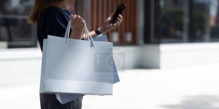Téléchargez les photos : Femme utilisant un smartphone tenant sac à provisions Black Friday tout en se tenant debout sur les escaliers avec le fond du centre commercial. - en image libre de droit