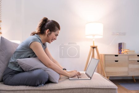 Téléchargez les photos : Jeune femme asiatique en utilisant ordinateur portable PC sur canapé se détendre surfer sur le net à la maison. - en image libre de droit