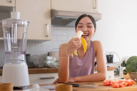 Téléchargez les photos : Jeune asiatique sain belle femme avec des vêtements décontractés est sourire et manger des fruits à l'alimentation à la maison dans la cuisine en vacances. - en image libre de droit