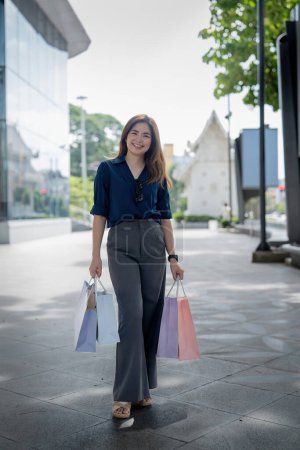 Foto de Feliz hermosa joven mujer con estilo asiático con bolsa de compras mientras camina cerca del centro comercial de vacaciones Viernes Negro. - Imagen libre de derechos