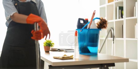 Photo for A cleaning woman is standing inside a building holding with a blue tank on the side facilities for tidying up in her hand. - Royalty Free Image