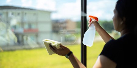 Téléchargez les photos : Jeune femme au foyer asiatique nettoyage vitre avec chiffons et pulvérisation de détergent, nettoyage maison le week-end. - en image libre de droit