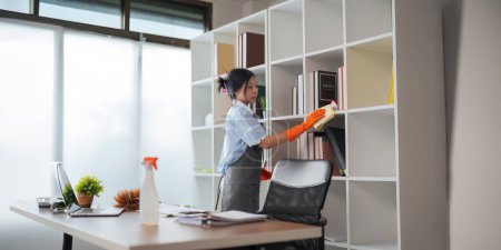 Foto de Mujer asiática limpiando en la sala de trabajo en casa. La joven limpiadora de limpieza usa un paño para limpiar el equipo para trabajar. concepto limpieza tareas domésticas limpieza. - Imagen libre de derechos