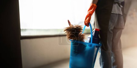 Photo for A cleaning woman is standing inside a building holding a blue bucket fulfilled with chemicals and facilities for tidying up in her hand. - Royalty Free Image