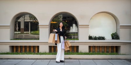 Téléchargez les photos : Heureuse belle jeune femme élégante asiatique avec sac à provisions tout en marchant près du centre commercial en vacances Black Friday. - en image libre de droit