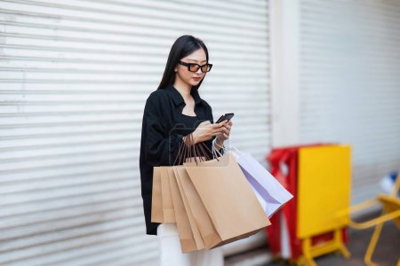 Foto de Mujer usando teléfono inteligente sosteniendo la bolsa de compras del Viernes Negro mientras está de pie en el lado con el fondo del centro comercial. - Imagen libre de derechos