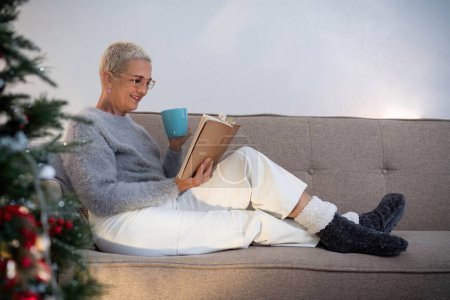 Téléchargez les photos : Femme âgée souriante avec une tasse de thé et un livre se détendre à côté d'un arbre de Noël décoré dans le salon. - en image libre de droit