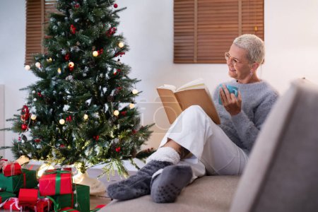 Téléchargez les photos : Femme âgée souriante avec une tasse de thé et un livre se détendre à côté d'un arbre de Noël décoré dans le salon. - en image libre de droit