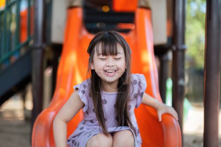 Photo for Smiling Asian little girl with fun sitting on slider at playground in the park. Education activity outdoor concept. - Royalty Free Image