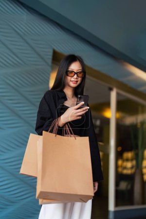 Téléchargez les photos : Femme utilisant un smartphone tenant Black Friday sac à provisions tout en se tenant sur le côté avec le fond du centre commercial. - en image libre de droit