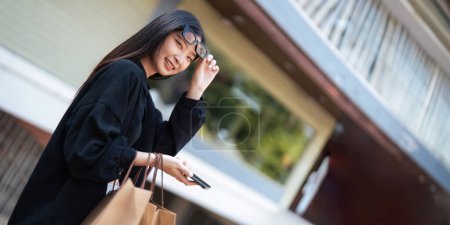 Photo for Woman using smartphone holding Black Friday shopping bag while standing on the side with the mall background. - Royalty Free Image