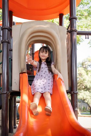 Téléchargez les photos : Asiatique petite fille jouer sur le curseur, Petite jolie fille est tout en jouant sur l'équipement de terrain de jeu dans le parc. - en image libre de droit