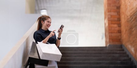Photo for Woman using smartphone holding Black Friday shopping bag while standing on the stairs with the mall background. - Royalty Free Image