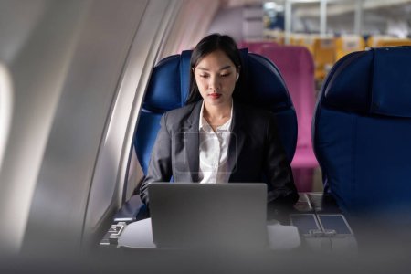 Foto de Hermosa mujer de negocios asiática sentada cerca de la ventana en el avión escuchar música en los auriculares mientras trabaja con el ordenador portátil. - Imagen libre de derechos