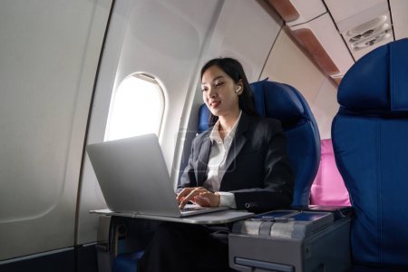 Foto de Hermosa mujer de negocios asiática sentada cerca de la ventana en el avión escuchar música en los auriculares mientras trabaja con el ordenador portátil. - Imagen libre de derechos