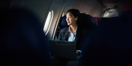 Téléchargez les photos : Femme d'affaires asiatique réussie, Femme d'affaires travaillant dans la cabine de l'avion pendant le vol sur ordinateur portable écoutant de la musique avec écouteurs. - en image libre de droit