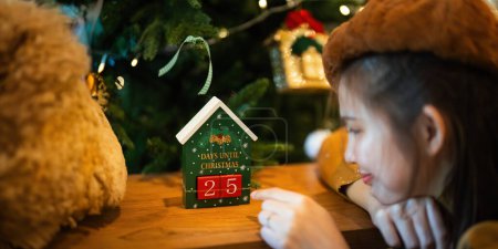 Téléchargez les photos : Une jeune femme s'assoit joyeusement en regardant un calendrier compter à rebours les jours jusqu'à Noël. concept temps de Noël. - en image libre de droit