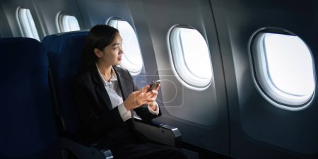 Foto de Exitosa mujer de negocios asiática, mujer de negocios que trabaja en avión en el ordenador portátil y mirando por la ventana junto con hablar por teléfono. - Imagen libre de derechos