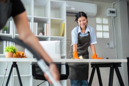 Foto de Mujer asiática limpiando en la sala de trabajo en casa. La joven limpiadora de limpieza usa un paño para limpiar el equipo para trabajar. concepto limpieza tareas domésticas limpieza. - Imagen libre de derechos
