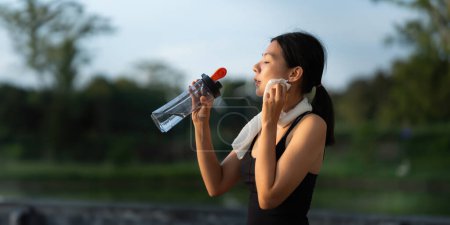 Foto de Hermosa joven asiática se detiene a beber agua y se limpia el sudor después de su carrera nocturna en el parque. - Imagen libre de derechos