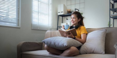 Téléchargez les photos : Joyeux jeune femme lire le livre sur le canapé à la maison. Lifestyle freelance se détendre dans le salon. style de vie concept de relaxation. - en image libre de droit