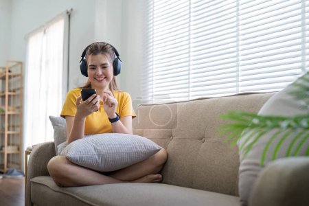 Photo for Young asian woman using mobile phone and earphones to listening to music or online chatting at home. - Royalty Free Image