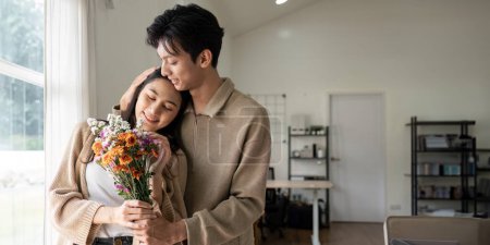 Foto de Romántico joven pareja asiática abrazando con la celebración de flores y sonriendo en la sala de estar en casa. enamorarse. Concepto Valentine. - Imagen libre de derechos