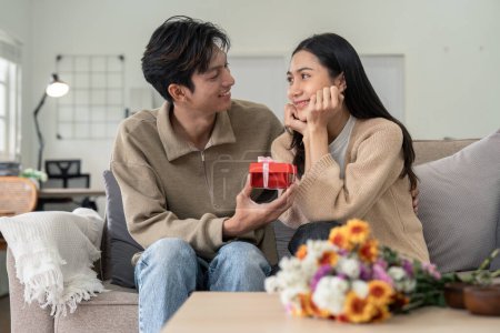 Téléchargez les photos : Romantique jeune couple asiatique embrassant donner cadeau dans le salon à la maison. Tomber amoureux. Concept de Valentin. - en image libre de droit