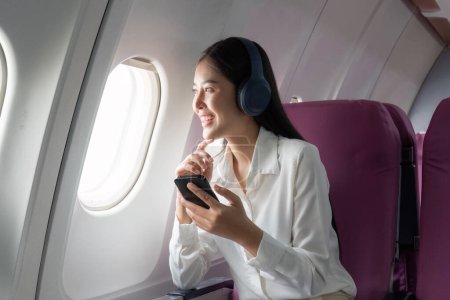 Téléchargez les photos : Femme asiatique joyeuse passager dans un casque regarder un film en ligne pendant le vol intercontinental en cabine de l'avion, voyageur heureux en utilisant une connexion wifi à bord. - en image libre de droit