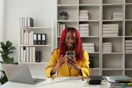 Téléchargez les photos : Femme d'affaires africaine utilisant l'application smartphone pour jouer à des jeux, faire des achats en ligne et commander la livraison tout en travaillant sur un ordinateur portable au bureau. - en image libre de droit